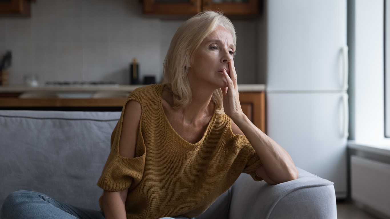 Woman sitting on a couch looking thoughtful and concerned, resting her hand on her face in a home setting.