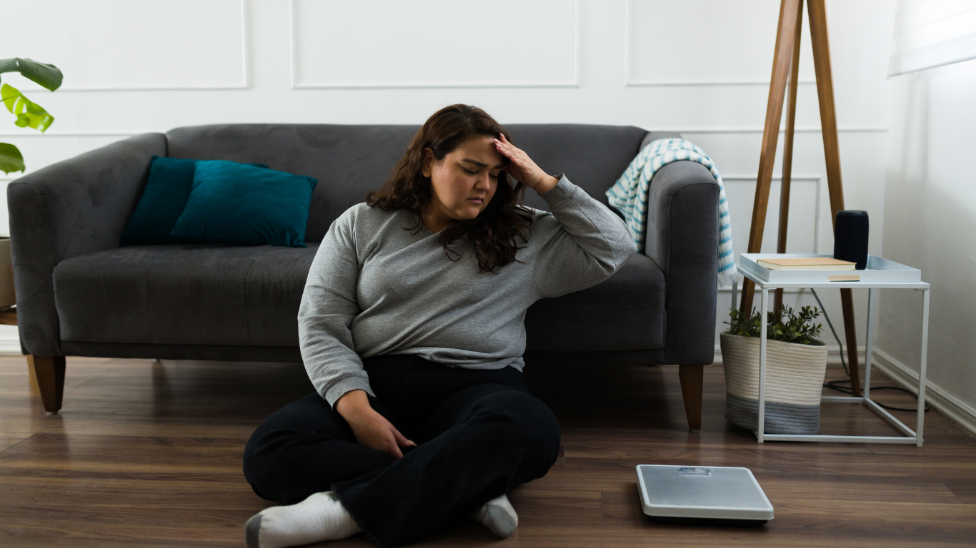 Woman sitting on floor looking fatigued next to a weighing scale at home